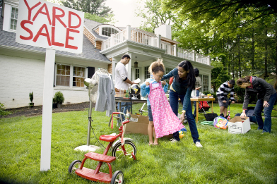 This photo shows a house with a sign that reads Yard Sale. There are people on the front lawn of the house, including a woman and a young girl looking at a girl’s pink dress that is for sale.