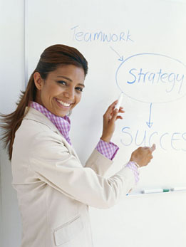This is a photo of a woman pointing at a whiteboard. The words teamwork, strategy, and success are written on the whiteboard.