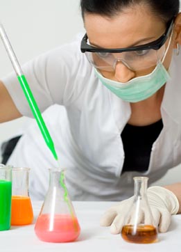 This photograph shows a female chemist wearing safety glasses, a lab coat, and a surgical mask while adding a solution to a beaker.