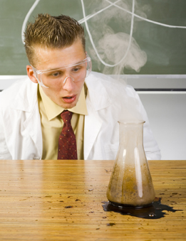 This is a photograph of a young man wearing safety glasses and a lab coat looking at a failed experiment where solution has been spilled from a beaker.