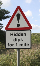 This is a photo of a triangular sign with an exclamation point in the middle. Below this sign is a another sign that reads “Hidden dips for 1 mile.”