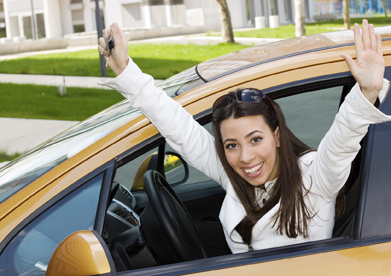 This shows a photo of a young woman with her hands raised while sitting in the driver’s seat of an orange car. She holds the keys to the car in her right hand.