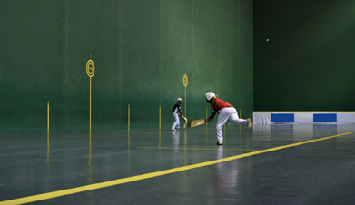 This is a photo of a jai alai court where a player in red has just thrown a ball off the front wall as a player in blue prepares to receive it.