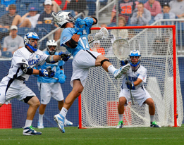 This shows a photo of an NCAA Lacrosse match between the Johns Hopkins Blue Jays and the Duke Blue Devils. A player is shooting on goal while a defender attempts to block the shot.