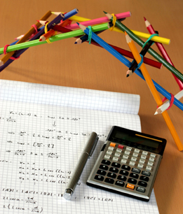 A model bridge formed from pencils and elastic bands is pictured on a desk by a calculator, a page of hand-written mathematics, and a pen.