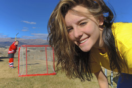 This photograph shows a female ball-hockey player with a male player swinging a hockey stick in the background.