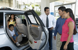 This is a picture of a car salesman showing an SUV to a family in a showroom.