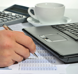 This photograph contains a laptop computer, coffee cup, calculator, and a person’s hand with a pen hovering over some stats on paper.