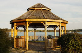 This photograph shows a wooden gazebo with a lake in the background. 