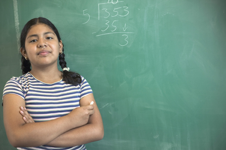 This photo shows a girl standing by blackboard. The blackboard shows the long division of 353 by 5. The quotient is 70 with a remainder of 3.