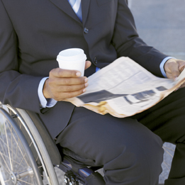 This photo shows a man in a wheelchair reading a newspaper and holding a coffee cup.
