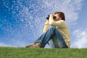 This is a photo of a young man sitting on some grass and looking at something with binoculars.