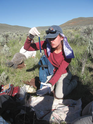 This photo is of a field technician in a scrub desert ecosystem in Utah. The technician sits on the ground with her equipment around her.