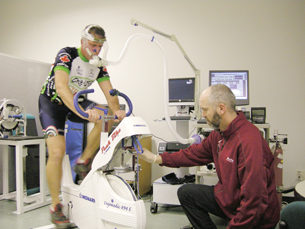 This photo is of a man riding a stationary bike in a medical lab.  He is wearing an apparatus around his mouth and nostrils that is hooked up to computers. A doctor is present to take readings.