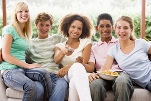 This picture shows diverse group of both male and female teenagers. They are all sitting on a couch. One girl is holding a bowl of chips. Another girl is pointing a remote control.