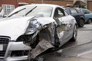 This photograph shows a silver two-door car that has been involved in a collision. The front fender and door on the driver’s side is badly damaged and the airbag has deployed.