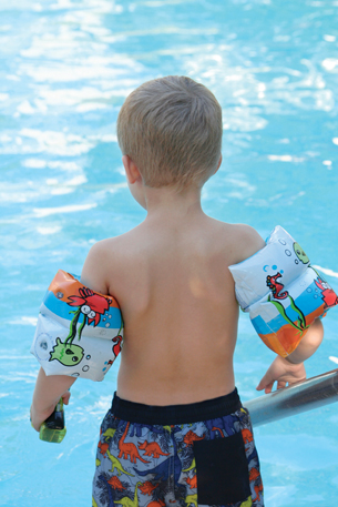 This photograph shows a young boy entering a swimming pool. The boy is wearing flotation bands on his arms.