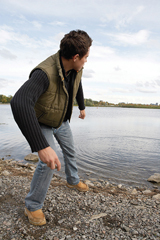 A photograph of a man throwing rocks into a lake in such a way that the rocks will skip along the water’s surface.