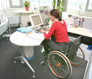 A photograph of a woman in a wheelchair working at a computer workstation.
