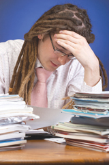This photograph shows a young man working at a desk. He is holding his head in one hand and looks overwhelmed by the stacks of work around him.