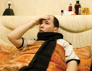 The photograph shows a young man in bed. He is bundled up in a blanket and is wearing a scarf around his neck. His face is flushed, and he has his hand to his forehead as if checking for a fever. When homeostasis cannot be achieved, a person becomes ill.