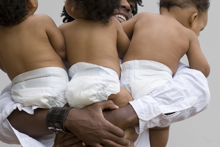 This photograph shows a dark-skinned adult holding three babies in diapers.