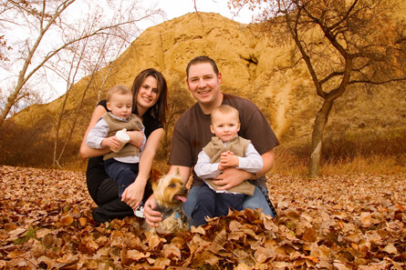 This family photograph shows a young couple, presumably the mother and father, each holding one of their twin boys, which appear to be around 1 year old. There is also a small brown dog in the photo.