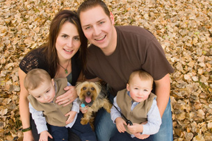 This photograph shows a family consisting of a young man, a young woman, infant twin boys, and a small-breed dog. It is posed as a family portrait, with all sitting close together and smiling toward the camera.