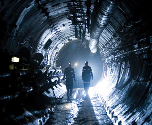 A photograph shows two men wearing coveralls and hard hats equipped with lamps walking in a dark underground tunnel. 