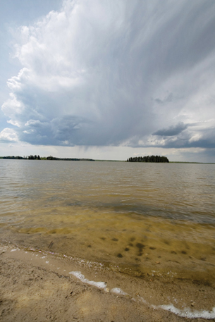 This photograph is from a beach alongside a lake. An island appears quite a distance from the shore. In the sky above the island is a large white cloud with a dark grey underside.