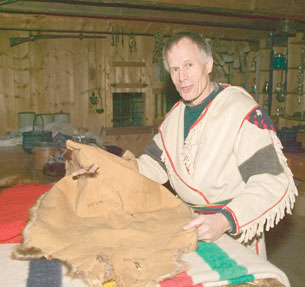 A photograph of a man examining a beaver pelt. The person is within a wooden building with many older technologies like oil lamps and power-loaded rifles. The man is wearing a wool tunic that is of a style worn by many Metis trappers in the late 1800s.