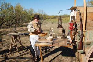 A man is preparing a meal on a wooden table at the back of a chuckwagon. Utensils being used are very rudimentary, bowls and simple spoons. Oil lanterns are hanging nearby.