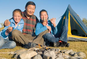 A elderly oriental man and his two granddaughters are roasting marshmallows by a campfire.  A nylon tent appears in the background.