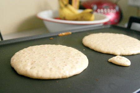 A photograph of two pancakes being cooked on a griddle. The surface of the griddle is black, indicating it has a coating. 