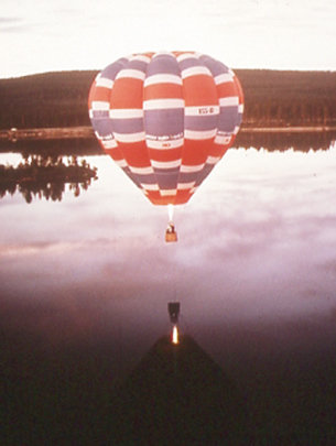 A photograph shows a hot air balloon floating over a lake.  The flame to heat the gases inside the balloon is visible in the space between the basket and the lower end of the balloon.