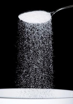 A photograph shows a spoon held above a white surface. Inside the spoon are many small white crystals. Some of the crystals are falling from the spoon.