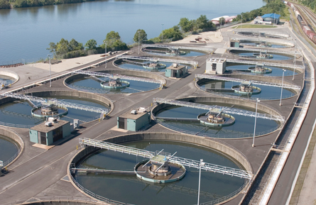 A photograph of a water treatment facility. The photograph shows 14 open circular tanks containing water. A narrow bridge extends across each tank. Within each tank is an arm-like extension from its centre.