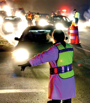 A photograph shows a night time scene in which police officers wearing reflective vests are directing cars to stop. Other police officers are talking with the drivers of the stopped vehicles.