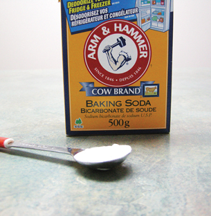 A photograph shows of a box of baking soda. In the foreground is a measuring spoon filled with a white powder.