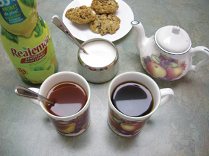 A photograph shows two cups of tea, a teapot, a sugar bowl, and a bottle of lemon juice. The colour of the tea inside the cup beside the lemon juice appears light brown. The colour of tea inside the other cup is a very dark brown-black in colour.