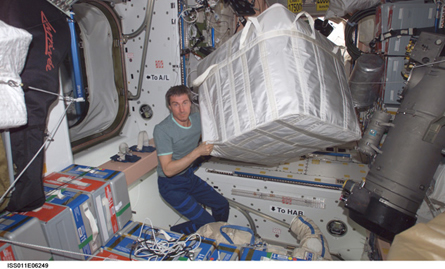 A photograph of an astronaut transferring a large container of supplies aboard the international space station. The container of supplies, which is as large as the astronaut, appears to float due to the low gravity conditions aboard the space station.