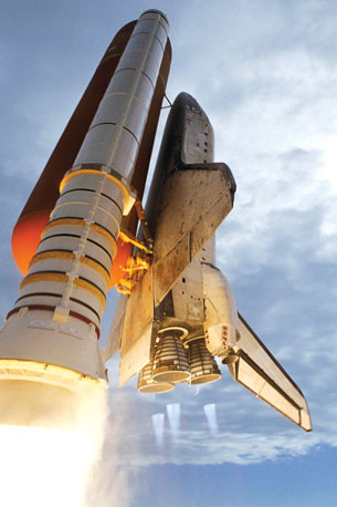A photograph of a space shuttle leaving the launch pad. Three blue-coloured flames are shown from the three rocket engines at the rear of the shuttle. Bright white flames are shown from the booster rockets attached to the large orange fuel tank on the underside of the shuttle.
