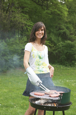A young woman is shown cooking hamburgers and hotdogs on an open charcoal barbecue.