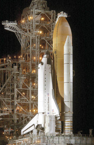 A photograph of a space shuttle on the launch pad.  The fuel tank for the main engines is a large orange-coloured cylinder that is attached to the underside of the shuttle. At the top of the fuel tank is a white ring that indicates where hydrogen and oxygen are added into the fuel tank.