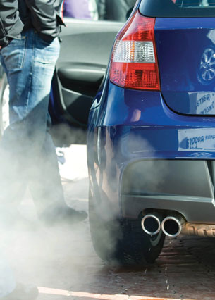 A photograph of a person standing beside the cloud of exhaust coming from an automobile tailpipe.