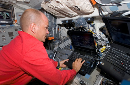 A photograph of an astronaut typing on the keyboard of a computer inside the crew compartment of a space shuttle.