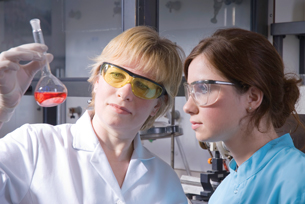 Two females are looking at a red liquid in a volumetric flask.