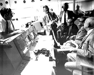 A photograph shows six men within the control room for the Apollo 13 mission having a conference. Each man has a concerned look on his face.