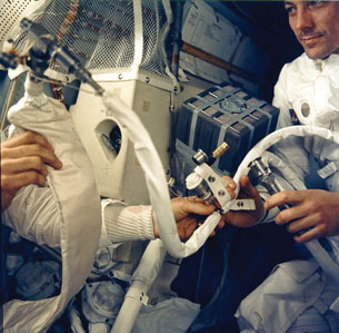 A photograph shows two astronauts floating inside the Apollo 13 spacecraft. The astronauts are making adjustments to a long white hose.