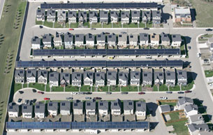 A photograph shows rows of houses, each with a solar panel on its roof. Behind each row of houses are garages with connected rooves. Along the upper surface of each roof are solar panels.